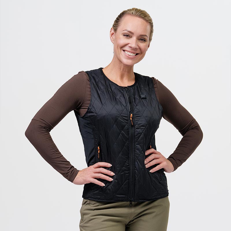 A woman wearing a black quilted heating vest from NordicHeat, standing with her hands at her sides and smiling, wearing a brown long-sleeved undershirt. The background is bright.