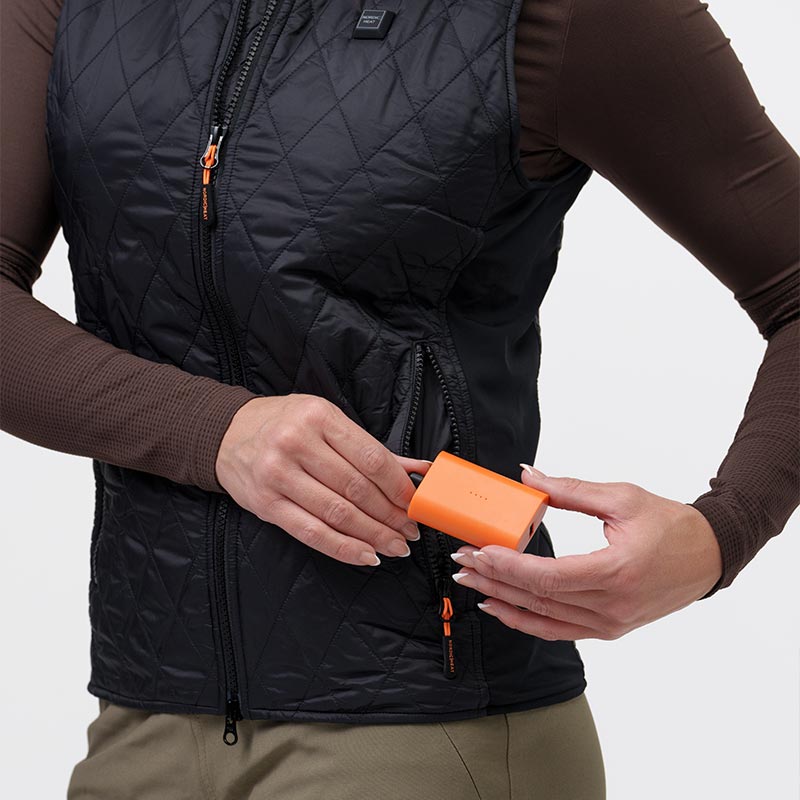 A person inserts an orange battery into the pocket of a black quilted jacket from NordicHeat. The jacket is worn over a brown sweater.