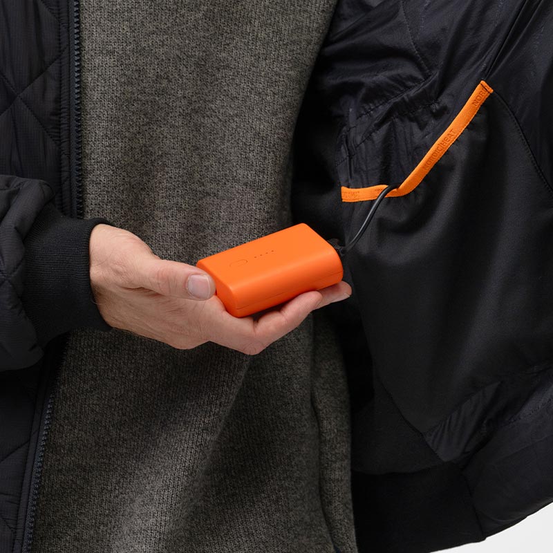 A hand holds an orange battery connected to a black piece of outerwear with NordicHeat branding on the inside.