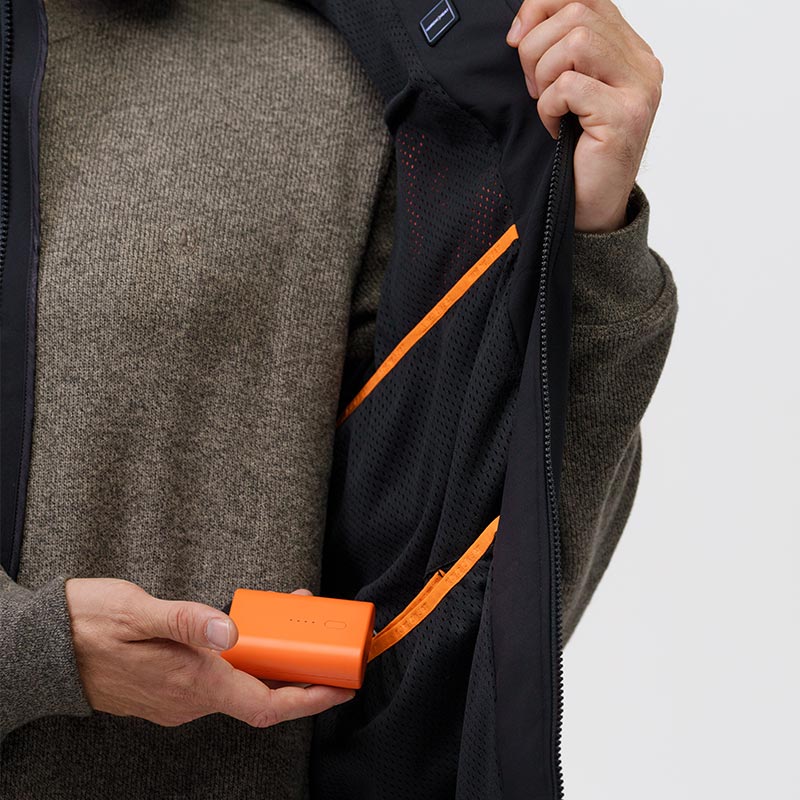 A person is holding an orange battery that plugs into a pocket in a jacket lined with black and orange details.