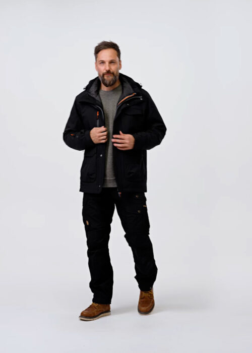 Man wearing black NordicHeat thermal clothing stands in a neutral studio setting. He has brownish boots, a gray shirt and a neat beard, with calm body language towards the camera.