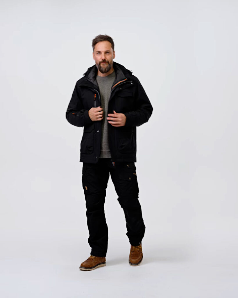 Man wearing black NordicHeat thermal clothing stands in a neutral studio setting. He has brownish boots, a gray shirt and a neat beard, with calm body language towards the camera.