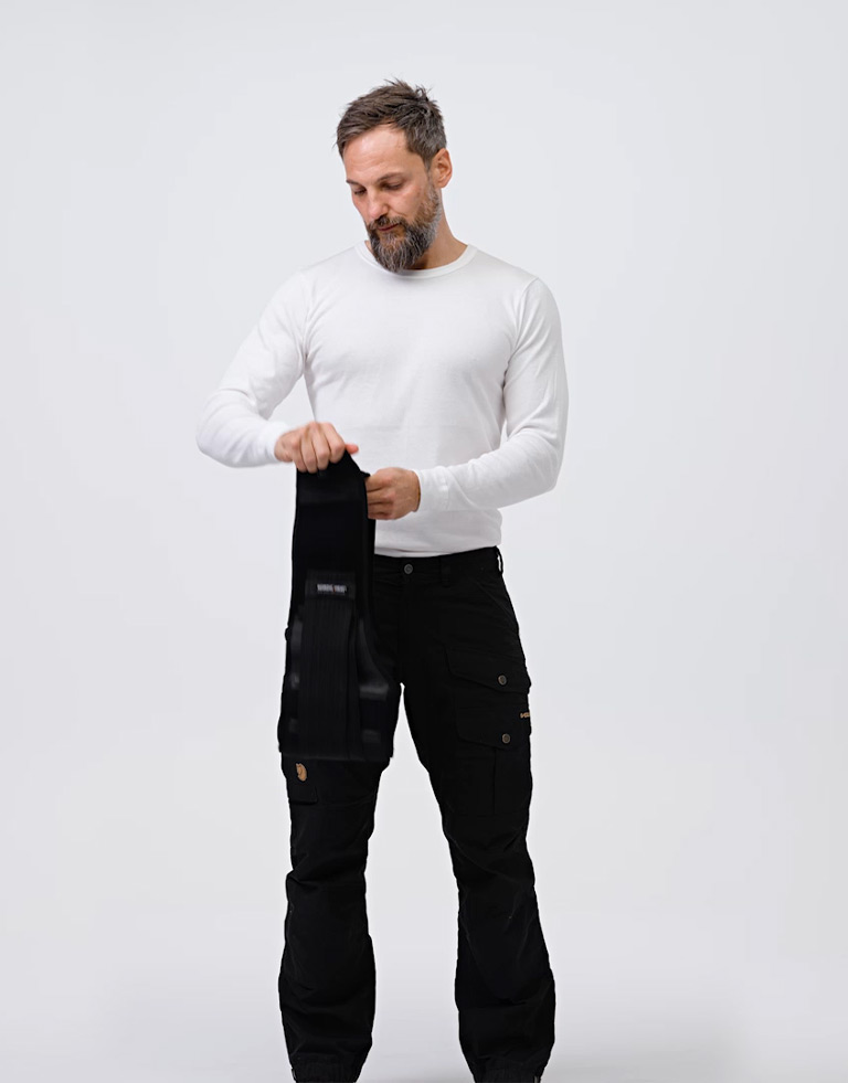 A man in a white sweater holds a piece of black NordicHeat thermal clothing against his body in a simple, indoor setting.