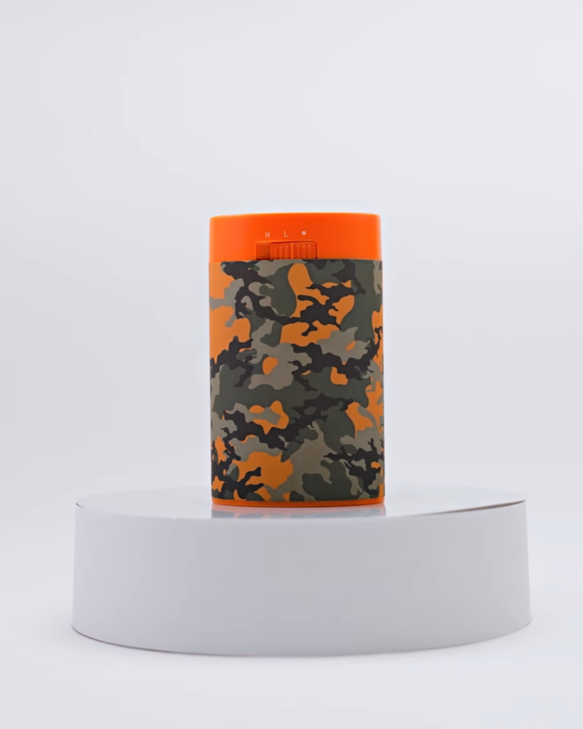 Cylindrical unit with camouflage print and orange top with settings marked as "H", "L", placed on a round white platform in a studio-lit environment.