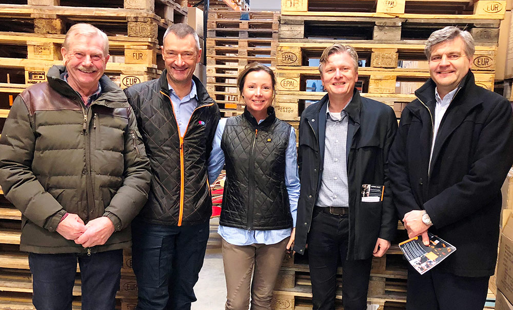 Five people in different NordicHeat warm jackets stand in a warehouse with pallets in the background. One person is holding a brochure.
