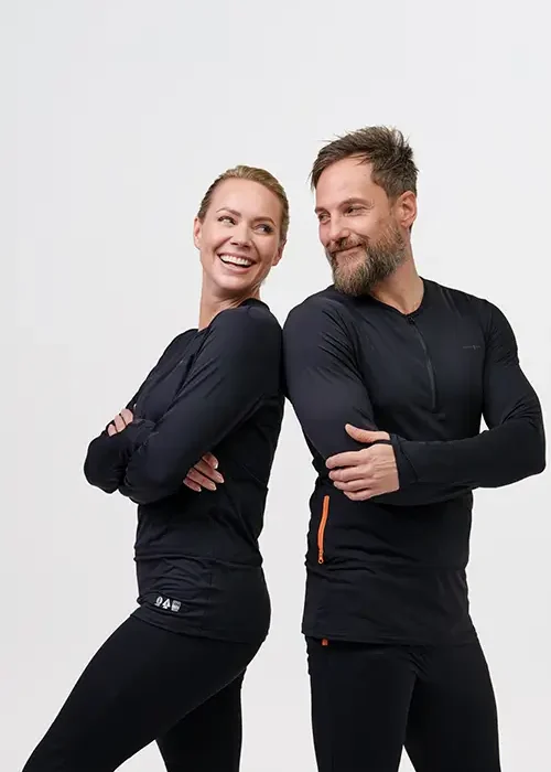 Two people wearing black NordicHeat warm clothing stand back to back and smile, against a neutral background, with a focus on comfort and functionality.