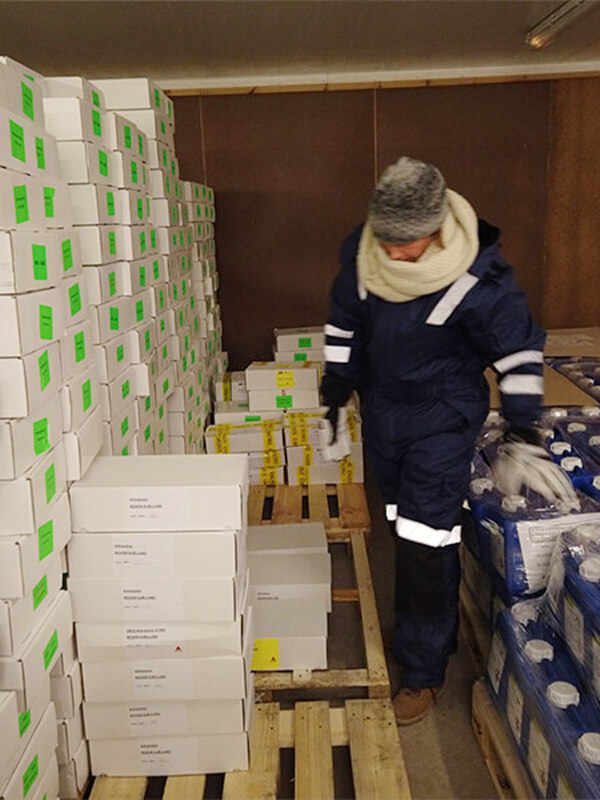 Person wearing NordicHeat warm clothing moves between stacked boxes in a cold room, doing warehouse work. Warm insulation products enable an active lifestyle in cold environments. Visible text: [no visible text].