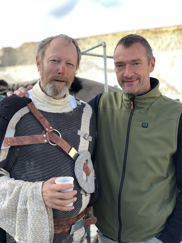 Two men stand together, one in armor, the other in a Nordic Heat vest, highlighting the functionality of thermal clothing. The background suggests a historical or active lifestyle.