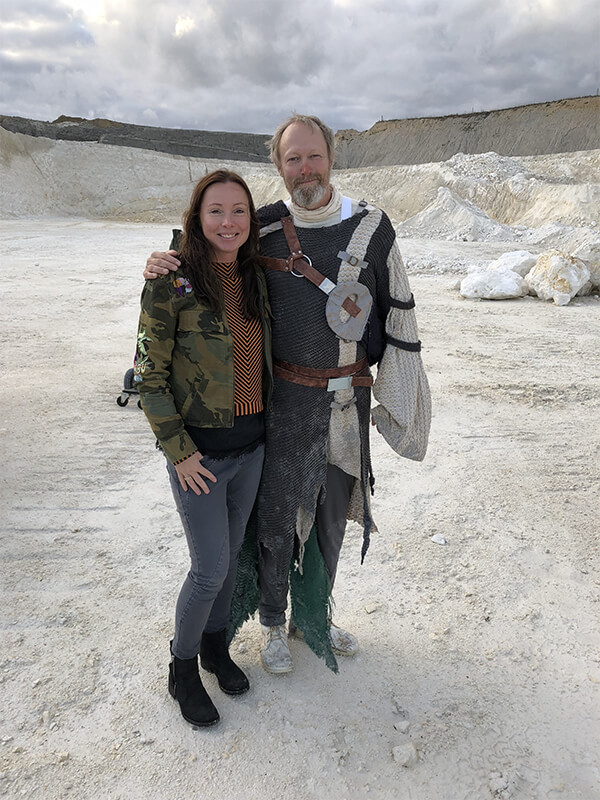 Two people are standing in a gravel area. The person on the left is wearing a warm jacket from NordicHeat, suitable for an active lifestyle. No visible text.