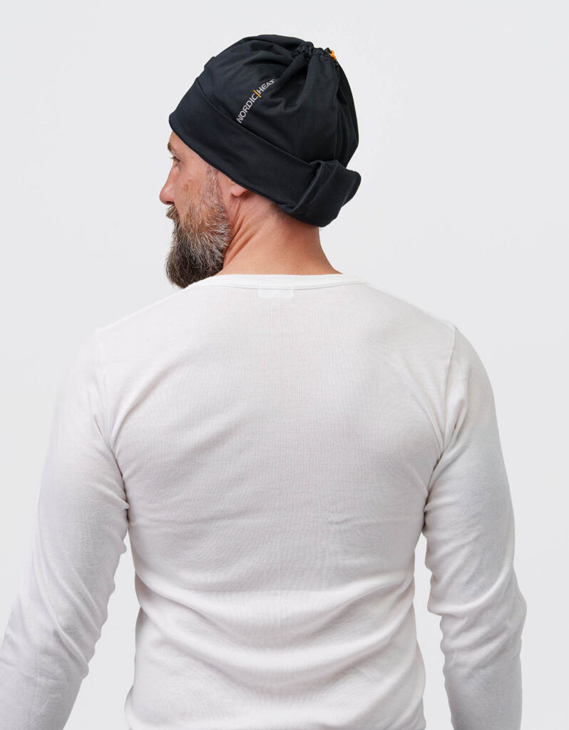 Person wears a black NordicHeat beanie that promotes warmth and comfort for active lifestyles. Text: "NORDICHEAT". Casual context focusing on practical clothing in a cool environment.