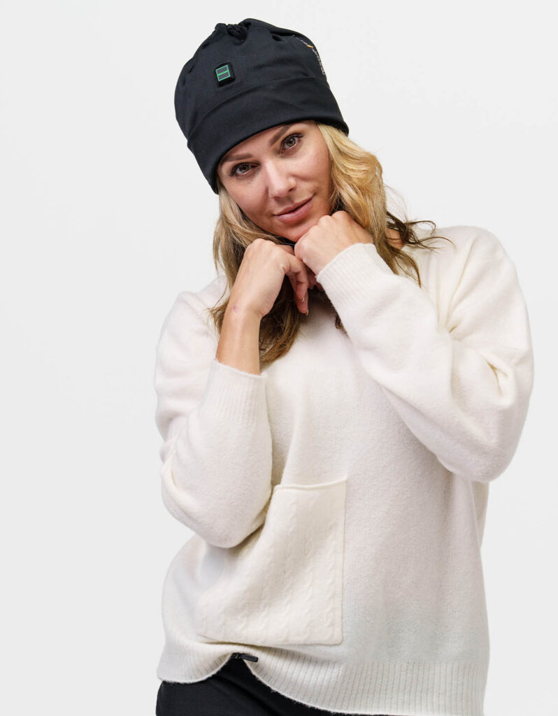 Person wears black NordicHeat warming beanie suitable for an active lifestyle. The cap has a green logo on the front. The person is dressed in a light sweater.