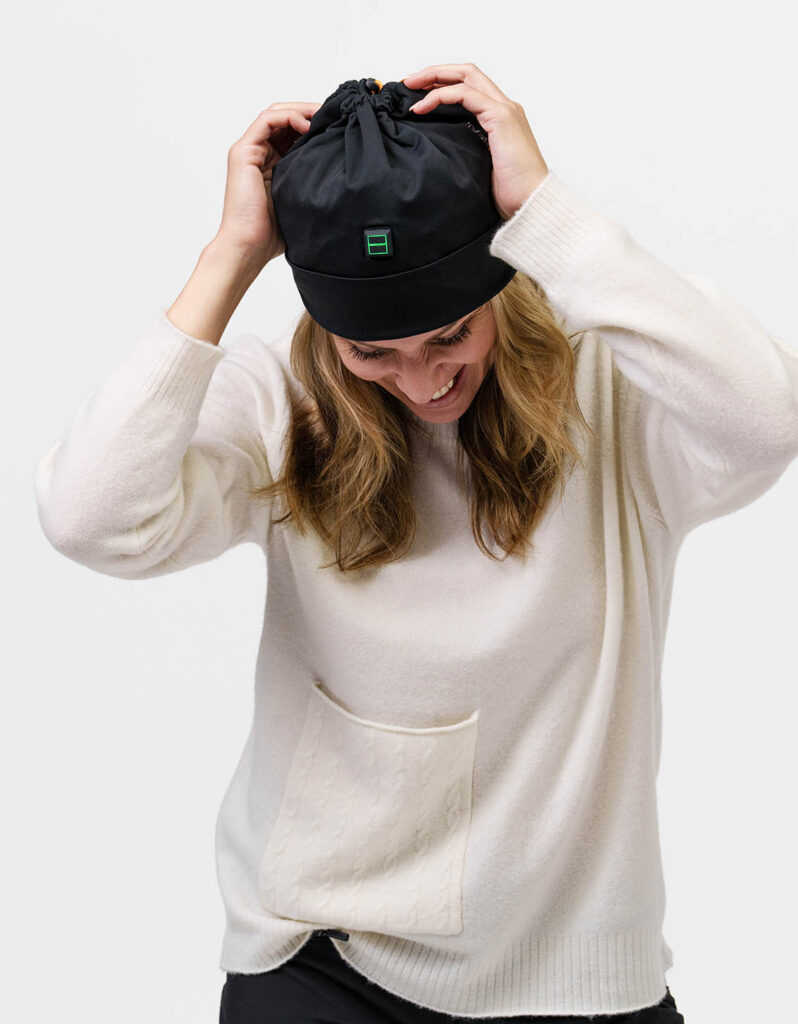 Person adjusts black NordicHeat beanie with heating panel, dressed in white sweater, symbolizing active lifestyle and practical winter clothing.