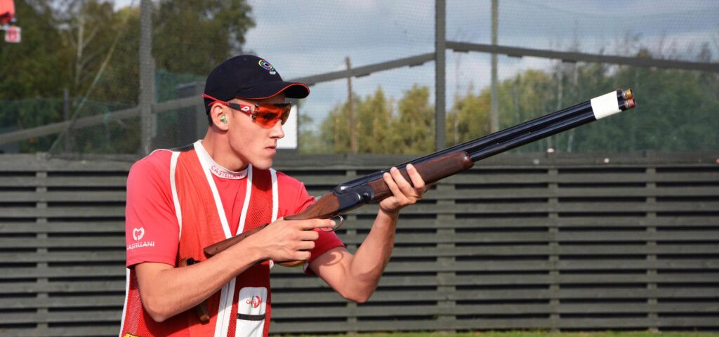 A person wearing NordicHeat thermal clothing takes aim with a sport shooter in an outdoor shooting competition. The heat helps maintain focus and performance. Visible text: "CASTELLANI".
