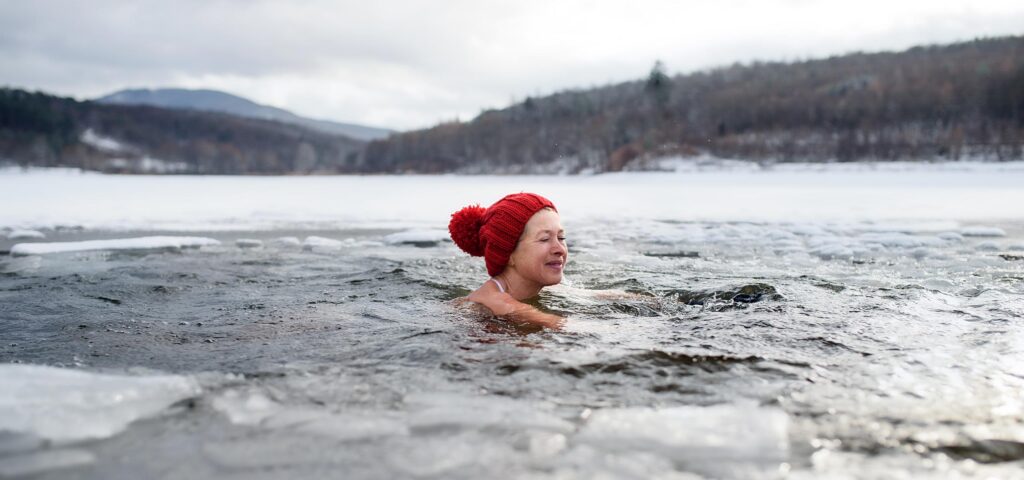 A person swimming in icy water wearing a red cap, surrounded by snowy landscape, symbolizes active lifestyles and the need for warmth products like NordicHeat's innovative warm clothing. No visible text.